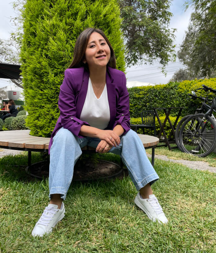 Gina Gonzáles sentada en un banco en un jardín, sonriendo a cámara con las manos curzadas.